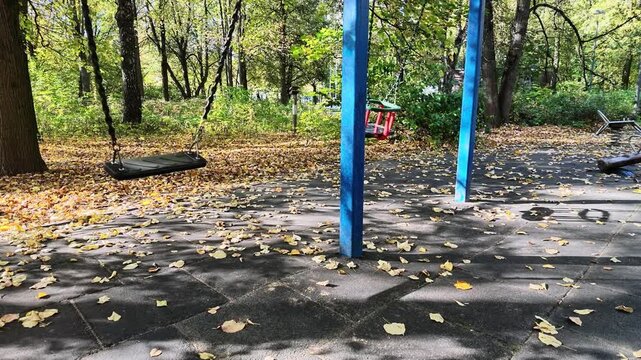 Empty swings sway on a playground covered with fallen leaves in a sunny autumn park.