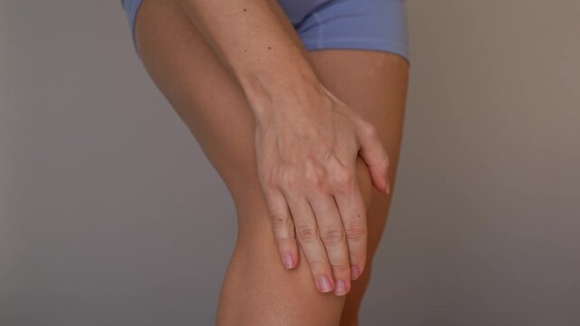 Young woman rubs ointment on painful knee, treating workout injury, joint pain, muscle soreness, and inflammation on gray background