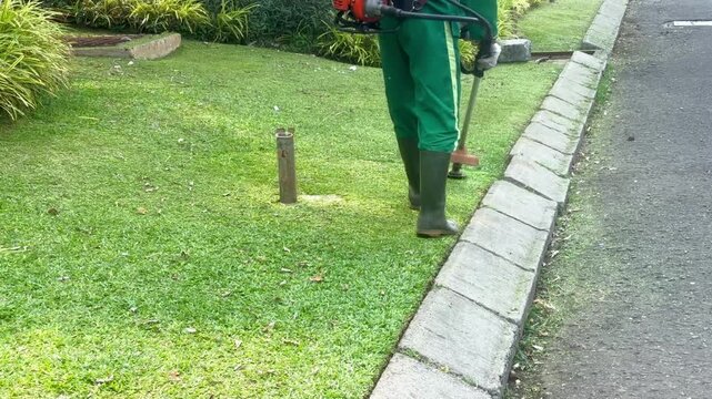 Worker mowing tall grass with electric or petrol lawn trimmer in city park or backyard. Gardening care tools and equipment. Process of lawn trimming with hand mower