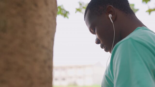 Man wears white earphones while standing near large brown textured object possibly outdoors with blurred green leaves and distant buildings. Soft natural lighting creates calm atmosphere