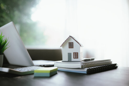 Small house model on stacked notebooks beside a laptop, representing real estate, home ownership, property investment, and housing planning.