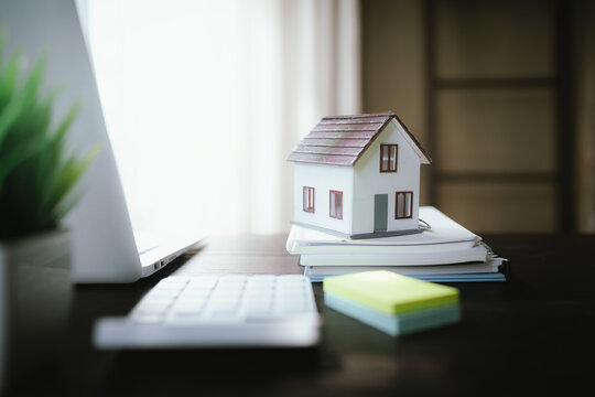 Small house model on stacked notebooks beside a laptop, representing real estate, home ownership, property investment, and housing planning.
