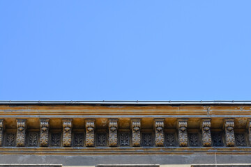 A scenic low angle view captures an old weathered building cornice featuring decorative stone corbels and carved relief panels beneath a clear blue sky. © nahhan
