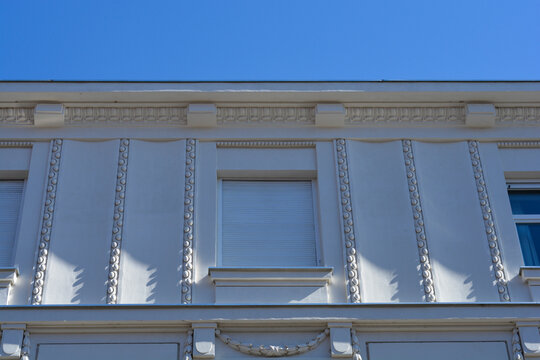 A detailed low angle view captures a white historic building facade featuring a window with closed rollers shutters and vertical wavy relief bands under a clear blue sky.