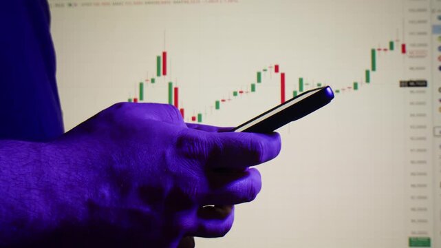 Phone in hand in front of stock market chart, close up of trader using smartphone trading app with candlestick graph on monitor background, investing online