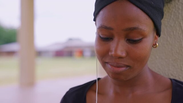 Close up shows young adult female subject in casual attire and gold hoop earrings. Woman wears white earphones against her ears while positioned next to outdoor structure with soft daylight