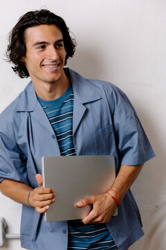 Young man in casual attire holding a laptop, looking cheerfully away.