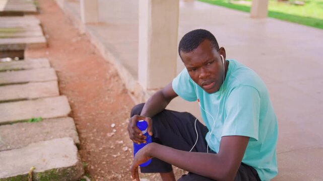 Young man sits on concrete step beside dirt path holding blue water bottle and wearing earphones. background includes columns with grass visible beyond them