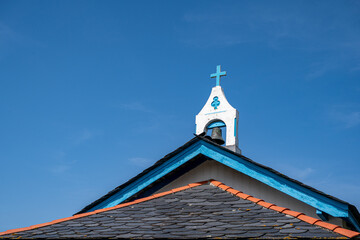 Church steeple with a blue cross stands against a clear blue sky. Red-tiled roof slopes upward toward the white bell tower. The scene exudes calmness and spiritual serenity. Ideal for themes of faith © AlbertoRodriguez