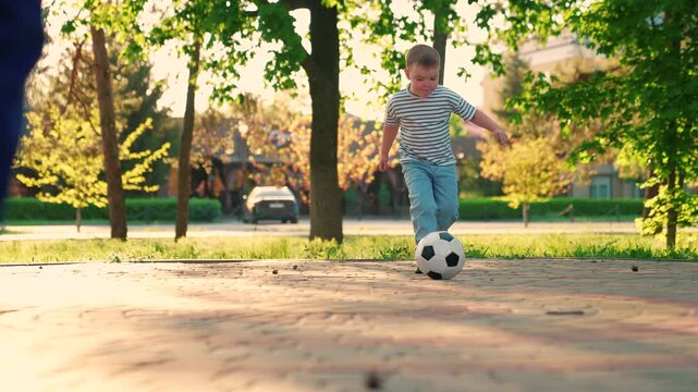 Child kicks football, dad son play soccer. Child kicks soccer ball and falls, kid laughs. Happy family plays football on weekends. Dad kid football in park. Family sports soccer team plays outdoors