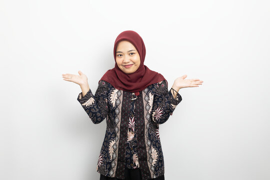 Asian Muslim woman in batik shirt and hijab shrugging shoulders isolated on white background. Ideal for don't know, doubt, choice, questioning, and problem concepts.