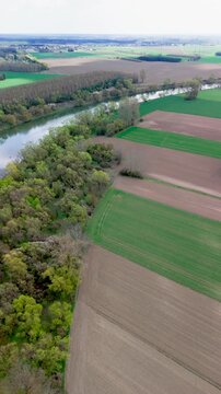 Vertical aerial view showing a river edged by thick green woodland beside striped farm fields. Plowed brown parcels alternate with young crops toward the hazy horizon, concept of balance between natu