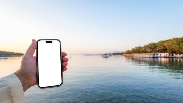 Hand holding a smartphone with a blank white screen against a serene coastal sunset landscape