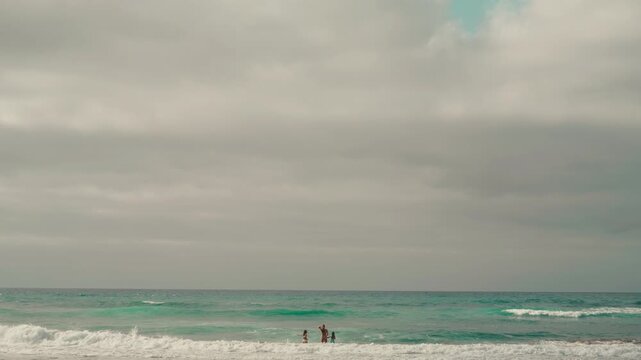 Coastal waves swirls as they run to sandy shore, gray gloomy sky hangs over beach of Gran Canaria, children play jumping in waves, slow motion. Seascape with breakers and gray clouds, Canary Islands