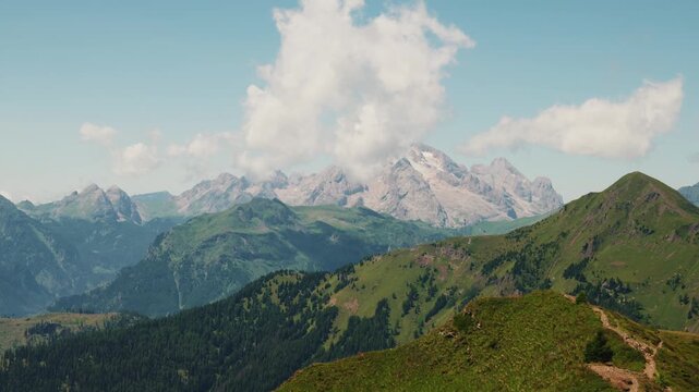 Wide landscape shot of high mountain range, glacier and green alpine meadows with hiking trail in Dolomites, Italy. Dramatic view of Marmolada glacier and green grassy ridges under cumulus cloud