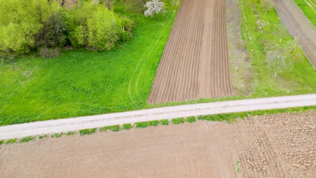 Narrow paved country road cuts across plowed brown plots and bright green grass fields with a hedge in the corner, aerial view of rural land parcels, concept of infrastructure, farm access and land p