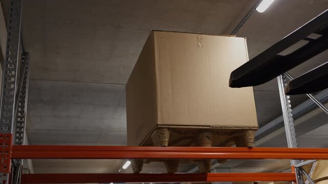 Forklift lowering a wooden pallet with a large cardboard box from a high storage rack inside a logistics warehouse