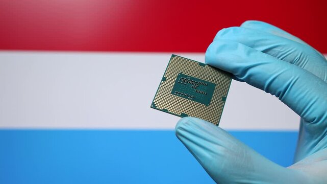 Close up view of hand in blue glove holding CPU microchip. Luxembourgish flag in blurred background. Concept of semiconductor shortage and artificial intelligence development in Luxembourg.