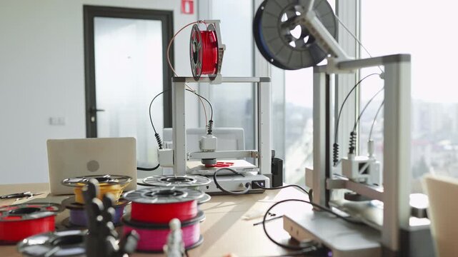 Multiple 3D printers and spools of filament are set up on a desk near a window, ready for production