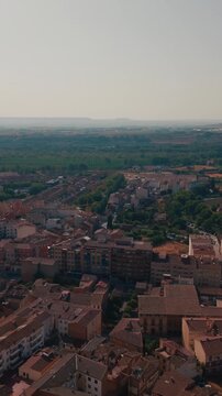 High angle pan view captures Monzon townscape around Aragon region. Terracotta roofs cover residential neighborhood facing green farm fields under hazy horizon during bright sunny day in Spain