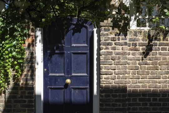 Blue wooden door with golden door knob in brick wall, partly in shade and surrounded by ivy, Chelsea, London, Great Britain, England, Great Britain