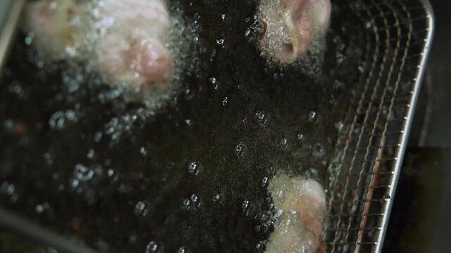 Vertical video. Frying bacon. Cooking food. Pork fat meat pieces preparing in bubbling hot oil inside professional deep fryer basket.