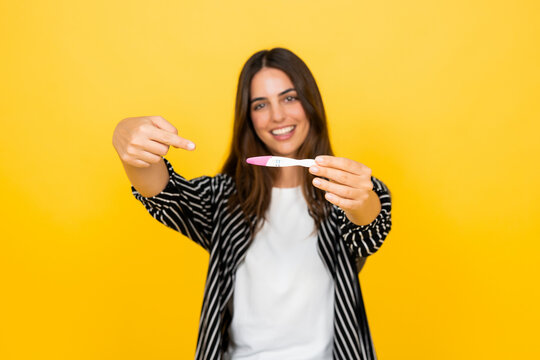 Young woman smiles and points to a positive pregnancy test on a yellow studio background. Her joyful reaction supports pregnancy announcement, fertility, family planning, motherhood and health.