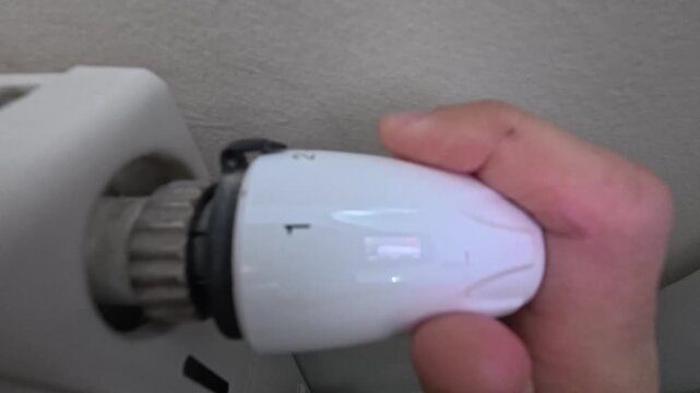 Hand turns radiator thermostat knob from numbered heat settings toward snowflake symbol. Thermostat control adjusts room heating, with fingers rotating white valve dial against textured wall.