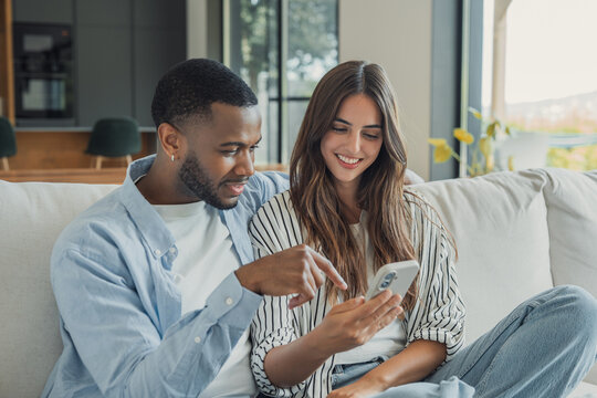 Interracial couple browse a smartphone together on a sofa at home while man points at the screen. Their shared smile illustrates mobile technology, online shopping, communication and connection.