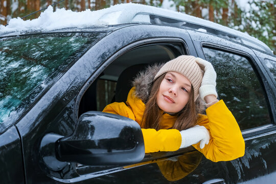 Woman driver taking a relaxing break during winter road trip, leaning from car window in snowy forest. Peaceful travel moment, rest and personal time concept.