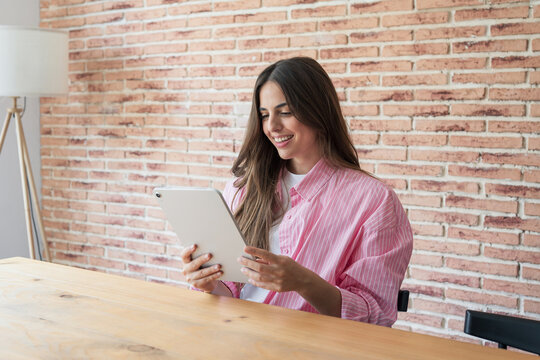 Young woman works on a tablet at home, reading the screen with a slight smile at a wooden desk. Casual remote work and freelance business technology concept in a bright brick-walled workspace indoors.
