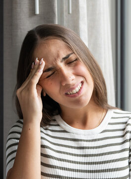 Headache affects woman holding her forehead beside a window at home. Eyes closed and a wince communicate migraine pain, tension, physical discomfort, and stress in softly lit indoor surroundings.