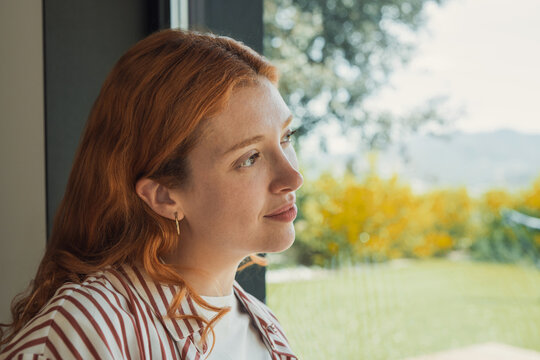 Woman gazes at nature through a home window in profile, framed by sunlit greenery. Her pensive expression and quiet indoor setting convey contemplation, solitude, mental wellness, and calm lifestyle.