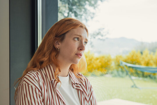 Redhead woman gazes outside from a home window in profile, framed by daylight and green foliage. Her melancholy expression supports themes of loneliness, anxiety, daydreaming, and mental wellness.