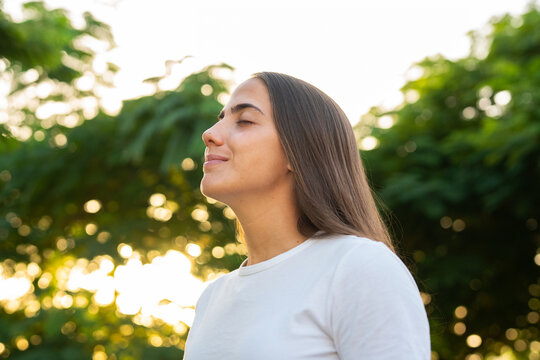 Woman breathes deeply outdoors with closed eyes, enjoying sunlit green trees and fresh air. Her calm relaxed pose supports wellness, mindfulness, mental health, relaxation and healthy living campaigns