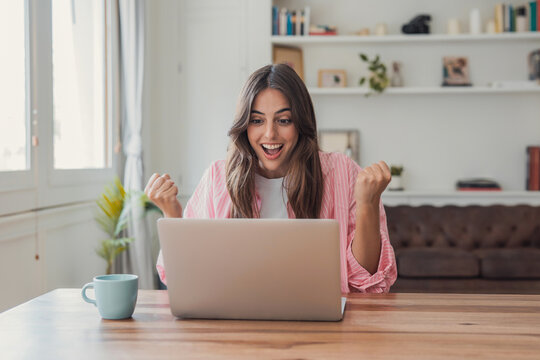 Woman celebrates a successful work result on her laptop at home, raising clenched fists with excitement. Remote office scene conveys freelance business achievement, good news and productivity.