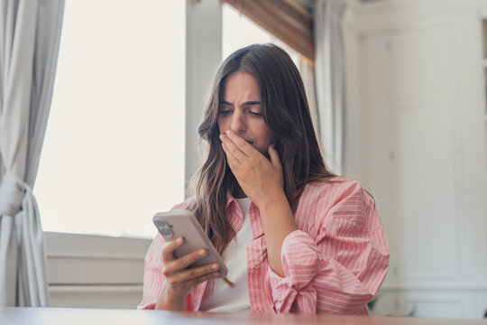 Woman covers her mouth while reading shocking news on a smartphone at home near a window, conveying surprise, concern, distress, bad news, and an unexpected digital notification.