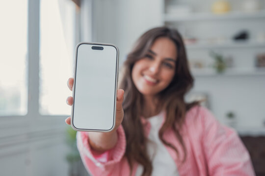 Woman holds a smartphone mockup with a blank white screen toward the camera, smiling warmly in a bright indoor room. Clean copy space supports app, website, advertising and interface design placement.