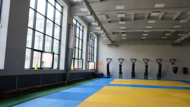 Empty indoor sports hall with blue and yellow training mats, large windows and wall-mounted exercise stations, suitable for gymnastics, martial arts, physical education, rehabilitation