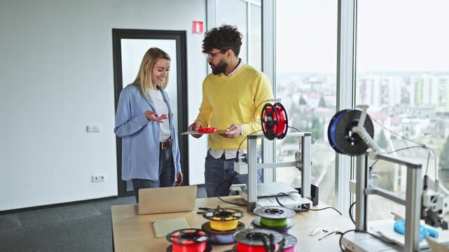 Two colleagues examine a 3D printed object next to a 3D printer and filament spools in a modern office setting