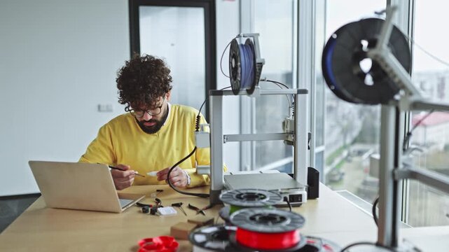 An Indian man with curly hair and glasses is meticulously lubricating the axles of a 3D printer during routine maintenance, surrounded by spools of filament and tools
