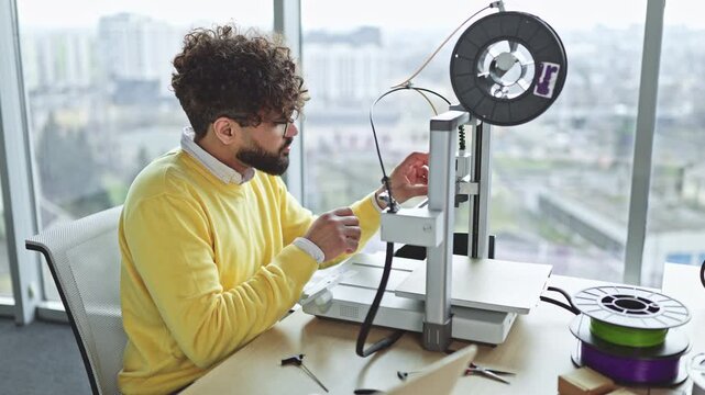 An Indian man with curly hair and glasses is focused on servicing the head of a 3D printer with a screwdriver, with spools of filament nearby