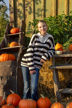 Young woman posing beside pumpkins and hay at an autumn harvest display, warm seasonal portrait with rustic decorations in golden sunlight.