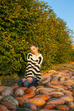 Young woman posing in a pumpkin patch during golden autumn sunset, seasonal outdoor portrait among colorful pumpkins, hay and green foliage.