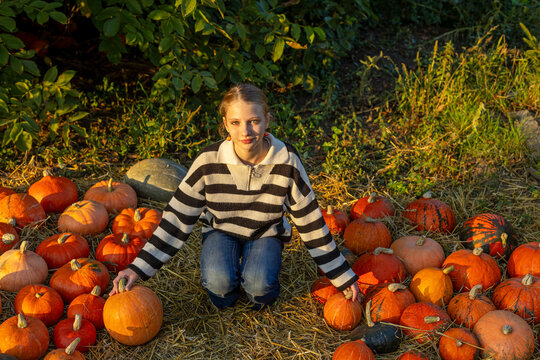Young woman kneeling among orange pumpkins at an autumn harvest display, warm sunset portrait surrounded by colorful seasonal gourds and hay.