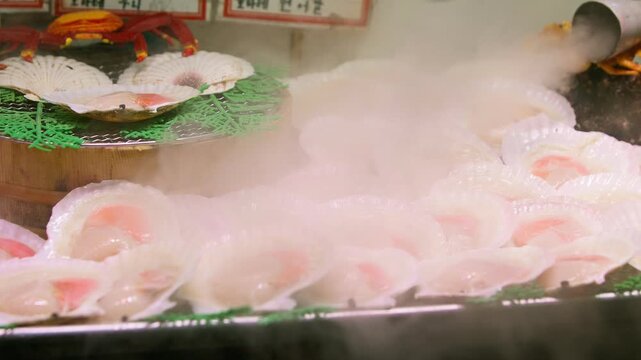 Captured in slow motion, pale scallops with orange roe rest inside ribbed shells on a heated display as wisps of steam drift across the rows, interrupted by a passerby in front of the market counter.