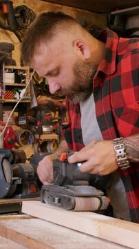 Vertical video. Carpenter occupation. Joinery process. Man smoothing board surface with belt sander in workshop interior with tools kit on shelves.