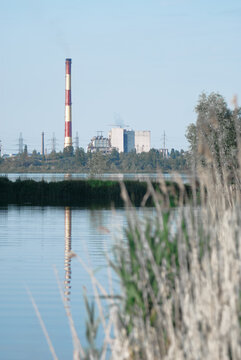 tall red-and-white smokestack rises behind an industrial power plant, its reflection mirrored in tranquil lake while reeds and trees frame the scene, highlighting contrast between industry and nature.