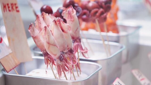 Captured in slow motion, pale squid bodies stand upright on wooden skewers inside metal trays, with tentacles hanging beneath the portions and darker octopus skewers visible behind them at a counter.