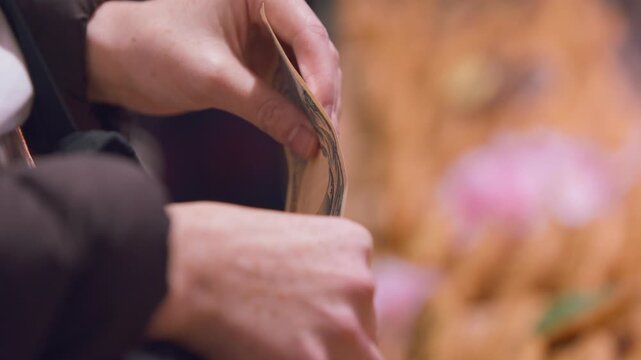 In slow motion, fingers separate and count yen banknotes in closeup beside a market food display, flexing the paper between both hands while orange and pink tones remain softly indistinct behind them.